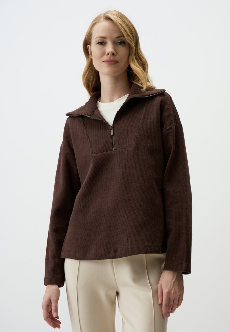 Pullover marrone a maniche lunghe con colletto a mezza zip e tessuto testurizzato. Presenta una cucitura frontale quadrata e una vestibilità rilassata. Indossato su pantaloni color crema.