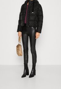 Veste matelassée noire à finition brillante, coupe courte, fermeture éclair, associée à des leggings en similicuir noir et des bottines noires à talons montant à la cheville.
