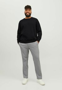 Pulover negru, pantaloni stripata gri deschis și adidași albi. Modelul stă cu mâinile în buzunare, pe un fundal simplu.