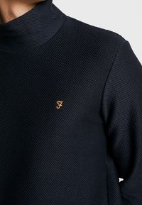  granatowy sweter z wysokim kołnierzem, teksturowany materiał, z małym pomarańczowym logo "F" po lewej stronie klatki piersiowej. Gładkie wykończenie, luźny krój.