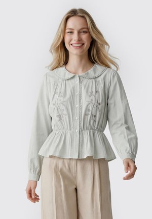 Smilende kvinde med langt blond hår iført en lysegrå knapbluse med blomsterbroderi og beige bukser, stående foran en hvid baggrund.