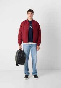 Red bomber jacket, navy sweatshirt, light blue jeans, and black sneakers. Holding a black structured bag. Simple design and casual fit.