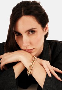 Bracciale a catena colore oro con anelli intrecciati, mostrato su un polso di carnagione chiara. Superficie liscia e lucida con anelli di diverse dimensioni.