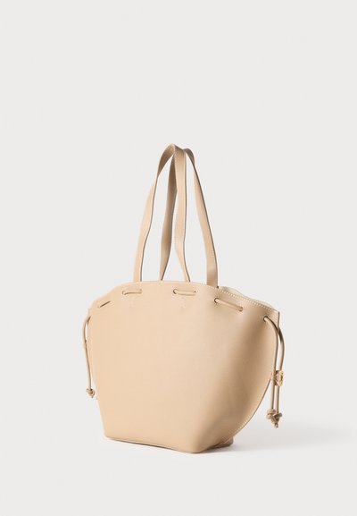 Τσάντα tote από μπεζ δέρμα με διπλές λαβές, στρογγυλεμένο σχήμα, κλείσιμο με κορδόνι στην κορυφή και αντίθετη ραφή. Λιτή υφή με ελάχιστα μεταλλικά στοιχεία.