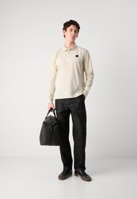 Lys beige langermet polo skjorte, svarte denimbukser, grå joggesko og en svart duffelbag. Minimalistisk design med subtile logodetaljer.