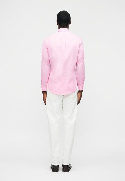 Άνδρας που στέκεται με την πλάτη γυρισμένη, φορώντας μακρυμάνικο ροζ πουκάμισο, λευκό παντελόνι και μαύρα παπούτσια, μπροστά σε ένα απλό λευκό φόντο.