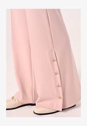 Lichtroze wijde broek van gladde stof, met gouden knoopdetails langs de zijnaden. Draagt erbij textuurrijke crèmekleurige en zwarte schoenen.