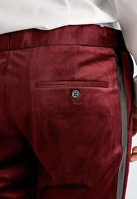 Close-up van bordeauxrode broek met een achterzak met knoop en een zwarte zijstreep, gedragen met een wit ingestopt overhemd.