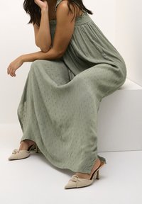 Robe maxi plissée verte avec des bretelles volantes et un tissu texturé. Assortie avec des escarpins beiges à bout pointu ornés d'un détail de nœud.