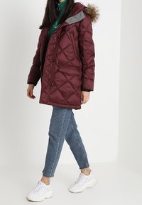 Mørkerød pufferjakke med diamantquiltning, grå hætte og pelsfor. Har for- knapper, lommer og en afslappet pasform. Kombineret med jeans og sneakers.
