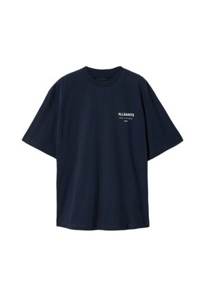 Jeune homme debout face à l'avant, portant un T-shirt AllSaints bleu marine, un pantalon beige et des baskets noires avec des accents blancs sur un fond uni.