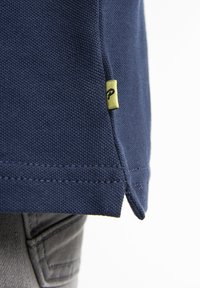 Marinblå polo med texturerad tyg, med en sidsspringa och en liten gul etikett med en svart logotyp på fållen.