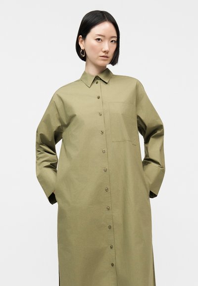 Γυναίκα με κοντά μαύρα μαλλιά που φοράει υπερμεγέθη ελιάς πράσινο φόρεμα-πουκάμισο με μακριά μανίκια και μπροστινή τσέπη, που στέκεται με τα χέρια στις τσέπες.