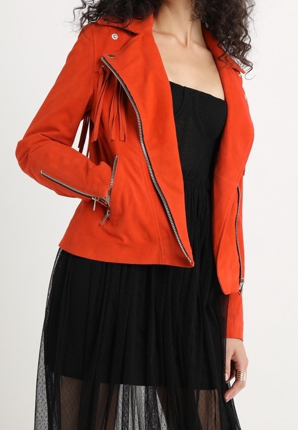 Veste de motard en daim orange avec des zips en argent, présentant des détails frangés sur les manches et un design ajusté, associée à une robe noire.