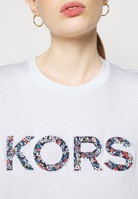 Bílý tričko s krátkým rukávem, na kterém je slovo "KORS" vyrobené z květinové látky s mnohobarevnými květy, orámované pevným bílým okrajem.