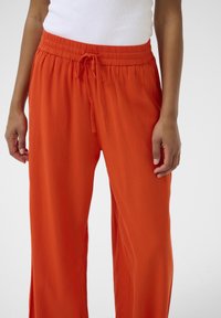 Oranžové široké kalhoty s elastickým pasem a šňůrkou, vyrobené z lehkého materiálu, které mají hladkou texturu a neformální design.