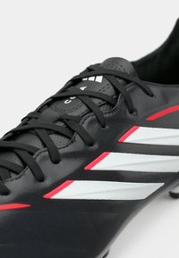 Μαύρο ποδοσφαιρικό παπούτσι με γυαλιστερό φινίρισμα, που έχει λευκές και κόκκινες ρίγες, μαύρα κορδόνια και υφασμάτινη υφή στην επάνω επιφάνεια.