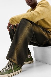 Person der sidder med benene bøjet, iført en gul fuzzy sweater, mørke bukser med brede ben, og grønne høj-top Converse sneakers.