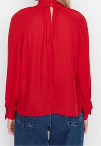 Red long-sleeve blouse with a high collar, back keyhole detail, and side slits at the cuffs. Smooth fabric with a flowing design.