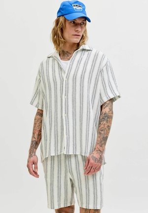 Mann mit Tätowierungen, der ein locker sitzendes weißes Hemd mit Knöpfen und Shorts mit schwarzen Streifen sowie eine knallblaue Baseballkappe trägt und nach rechts schaut.