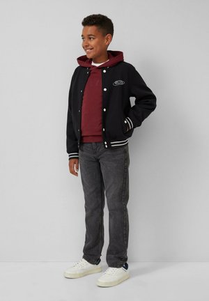 Ein lächelnder Junge steht in einer schwarzen Collegejacke, einem kastanienroten Hoodie, dunklen Jeans und weißen Sneakern vor einem schlichten grauen Hintergrund.