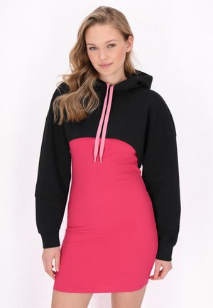 Junge Frau mit welligem blondem Haar trägt einen schwarzen kurzen Hoodie mit pinken Kordeln über einem eng anliegenden pinken Kleid.