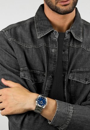 Orologio d'argento con quadrante blu, cinturino in acciaio inossidabile, indossato al polso. Lo sfondo mostra una giacca di jeans grigia con dettagli neri.