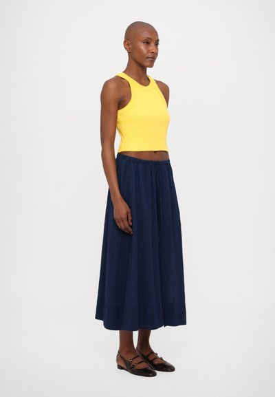 Geel cropped tanktop, marineblauwe vloeiende maxi rok met trekkoord taille, en bruine schoenen met banden. Soepele texturen, eenvoudig ontwerp.