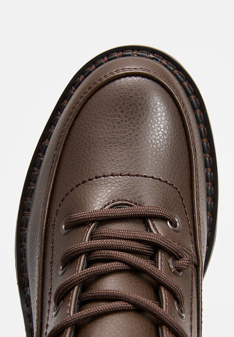 Braunes Leder-Schuhwerk mit strukturierter Oberfläche, runder Spitze und genähten Akzenten. Verfügt über braune Schnürsenkel und eine robuste Sohle mit einem dunklen Rand.