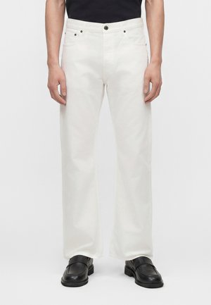 Άνδρας που στέκεται φορώντας λευκά τζιν παντελόνια με ίσια γραμμή και μαύρα δερμάτινα μοκασίνια σε απλό φόντο.