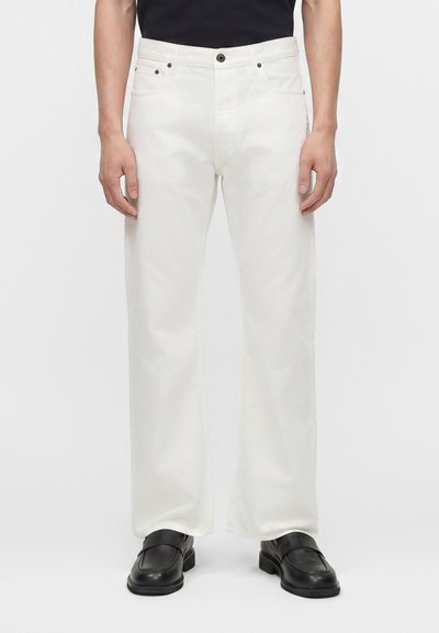 Homme debout portant un jean blanc droit et des mocassins en cuir noir sur un fond uni.
