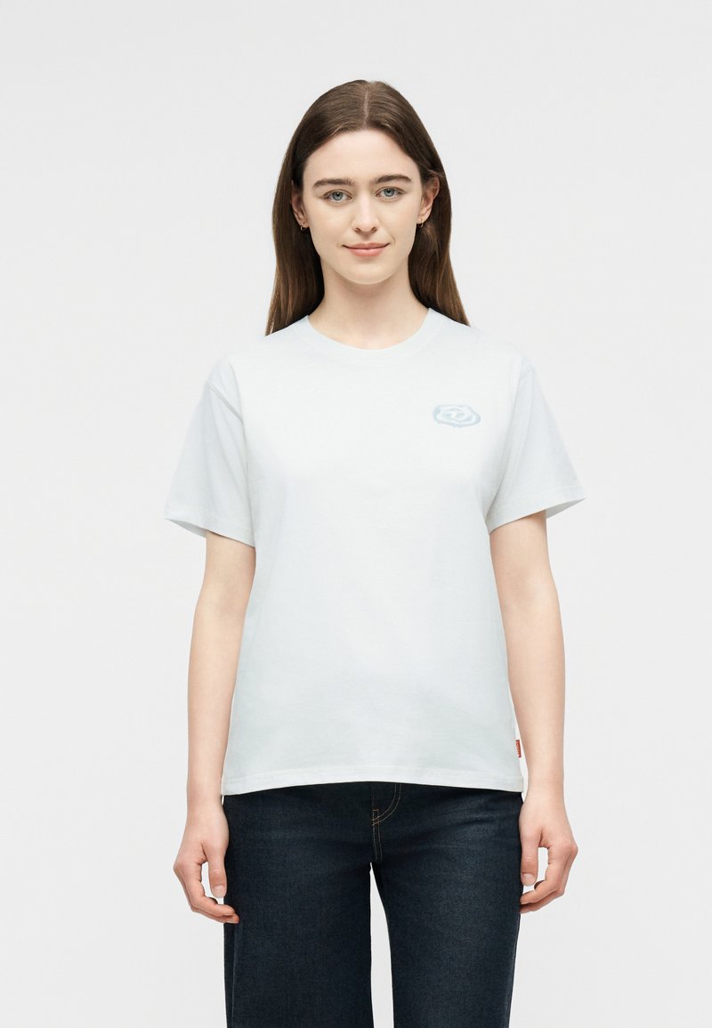 Νεαρή γυναίκα με ίσια καστανά μαλλιά, που φοράει απλό λευκό κοντομάνικο μπλουζάκι και σκοτεινό τζιν, στέκεται μπροστά σε ανοιχτό φόντο.