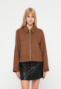 Καφέ σατινέ μπουφάν με φερμουάρ μπροστά, μυτερό γιακά και μακριά μανίκια. Συνδυασμένο με μαύρη γυαλιστερή δερμάτινη μίνι φούστα με κουμπιά.