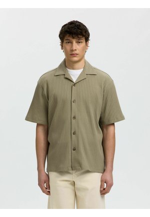 Młody mężczyzna ubrany w beżową prążkowaną koszulę na guziki z krótkim rękawem na białą koszulkę i jasne spodnie, stojący na jednolitym tle.