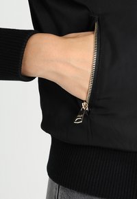 Giacca bomber nera con tasca con zip e hardware dorato, polsini a coste e tessuto opaco.