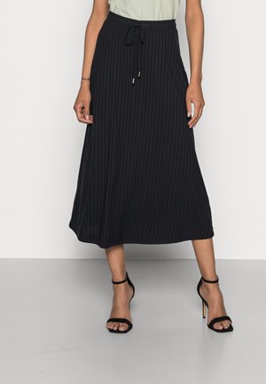 Femme portant une jupe mi-longue plissée noire avec une taille à cordon et des sandales à talons hauts noires, debout devant un fond uni.
