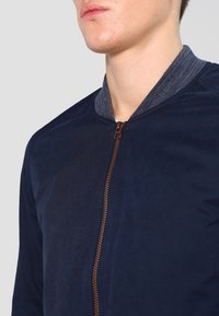 Marineblauw bomberjack met een gladde textuur, voorzien van een contrasterende grijze geribde kraag en een bruine rits. Close-up weergave die de details benadrukt.