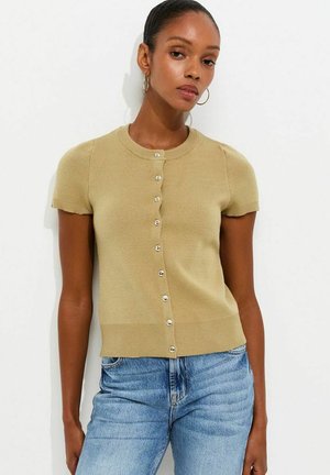 Vrouw draagt een beige, kort mouwen geknoopte cardigan en blauwe jeans, staand tegen een witte achtergrond, met haar naar achteren en hoepeloorbellen.