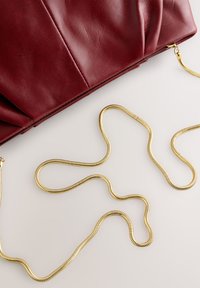 Bordeauxrode leren handtas met een geplooid ontwerp, vergezeld van een gouden kettingriem die in een golvend patroon op een lichte achtergrond ligt.