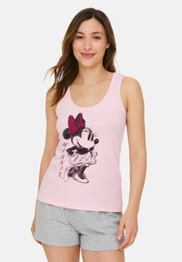 Donna che indossa una canottiera rosa con una grafica di Minnie Mouse e pantaloni grigi, in piedi e sorridente su uno sfondo bianco.