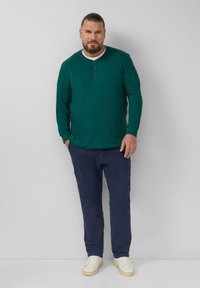 Długosleeve'owa zielona koszula henley z białym kołnierzykiem, dwoma guzikami i granatowymi spodniami cargo, zestawiona z białymi trampkami na jednolitym tle.