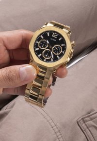 Aukso tonų laikrodis su oktaedriniu korpusu, juodu ciferblatu, sidabriniais akcentais ir chronografo funkcijomis. Metaliniai apyrankės elementai suteikia elegantišką išvaizdą.