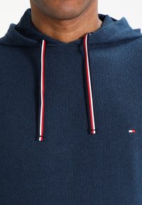 Felpa blu navy realizzata in tessuto testurizzato, con lacci rossi e bianchi, e un piccolo logo in basso a destra.