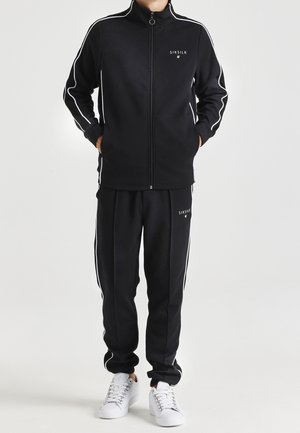 Osoba ubrana w czarny dres SikSilk z białymi obszyciami i białe trampki, stojąca z rękami w kieszeniach na jednolitym tle.