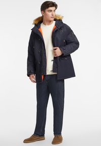 Parka aislante de marina con capucha forrada de piel, forro interior naranja, cierre de botones y cremallera, combinado con jeans oscuros y zapatos marrón claro.