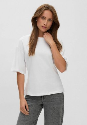 Jeune femme aux longs cheveux bruns portant un t-shirt blanc uni à manches courtes et un jean gris sur un fond clair.
