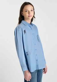 Light blue button-up shirt with a vertical stripe pattern. Features black embroidered lightning bolts and stars, single chest pocket, long sleeves.