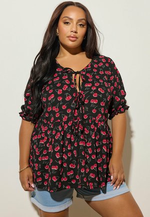 Donna che indossa una camicetta nera con stampa di ciliegie rosse e pantaloncini di jeans azzurri arrotolati, in piedi contro uno sfondo chiaro e semplice.