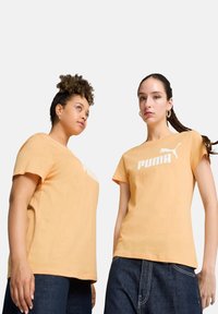 Oranžid puuvillased T-särgid lühikeste varrukatega ja valge PUMA logo. Ühel mudelil on lokkis juuksed ning teisel sirged juuksed, millel on punutis.