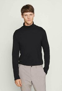 Young man with short brown hair wears black turtleneck and light gray pants, standing with one hand in pocket against a plain background.